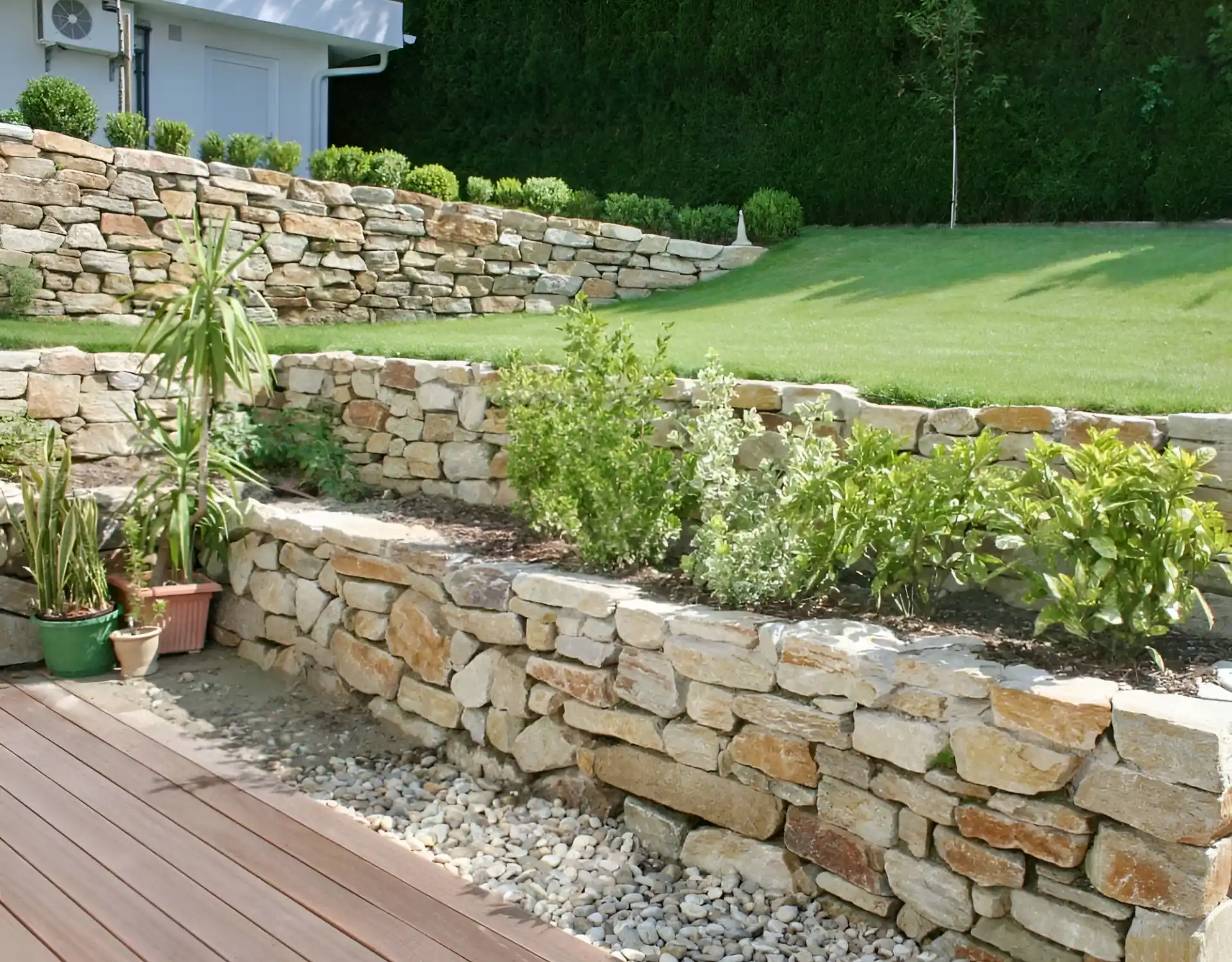 Mehrstufige Natursteinmauer mit Bepflanzung, Kiesstreifen und Holzterrasse in einem terrassierten Garten